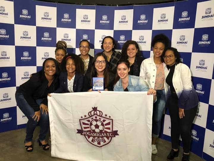 Mulheres segurando troféu e bandeira do time de futebol feminino Jogamigas