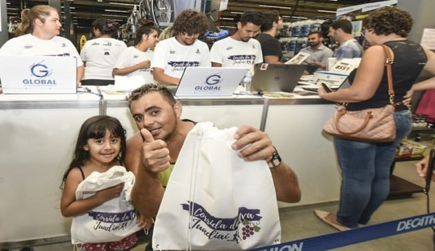Menina segurando sacola da festa da uva, à esquerda; homem com luzes loira no cabelo e regata verde abaixado ao lado da menina, também segurando uma sacola da festa da uva, à direita; ambos estão na frente do caixa com notebooks escrito: "Global"
