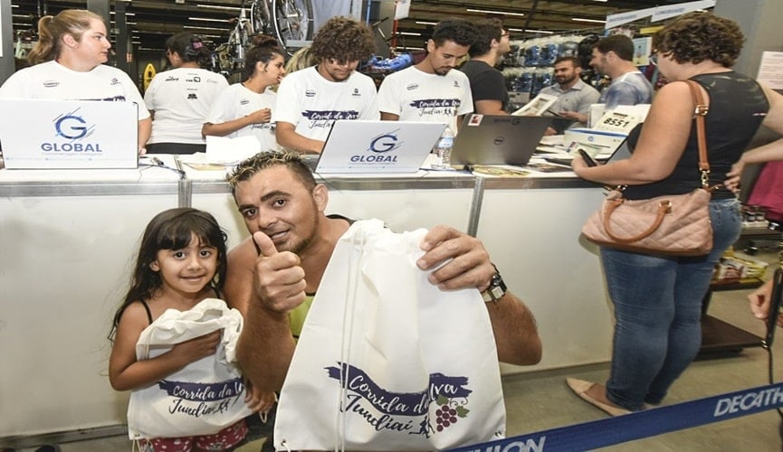Menina segurando sacola da festa da uva, à esquerda; homem com luzes loira no cabelo e regata verde abaixado ao lado da menina, também segurando uma sacola da festa da uva, à direita; ambos estão na frente do caixa com notebooks escrito: "Global"