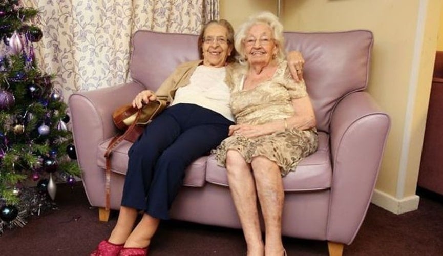 Idosas sentadas em sofá lilás, ao lado de uma árvores de natal, em uma sala amarela