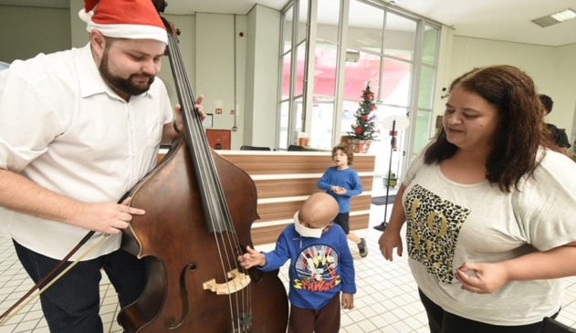 Criança com câncer tocando em instrumento de músico com gorro de Papai Noel, ao lado da mãe
