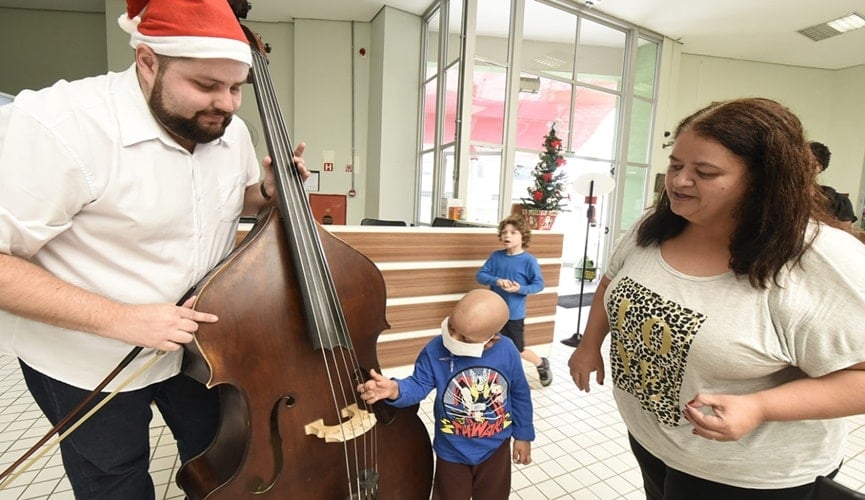 Criança com câncer tocando em instrumento de músico com gorro de Papai Noel, ao lado da mãe
