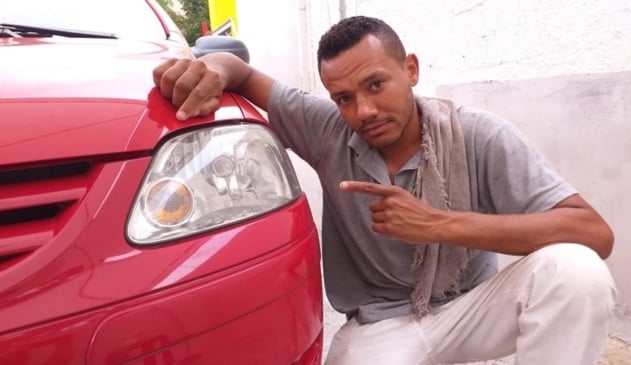 Homem negro ao lado de carro vermelho apontando para faról