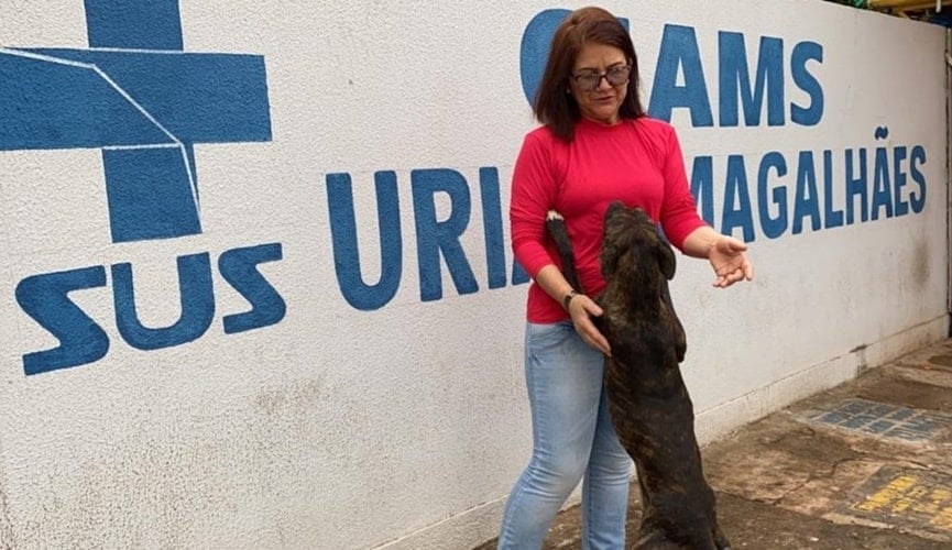 Mulher de meia idade vestida com blusa rosa abraçada com cachorro escuro na frente do muro da Unidade Básica de Saúde