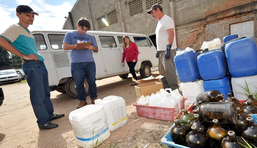 Foto de uma perua branca com uma mulher loira ao lado, três homens à frente; o da esquerda está observando uma pilha de lixo, o do centro está anotando, o da direita está recolhendo o lixo