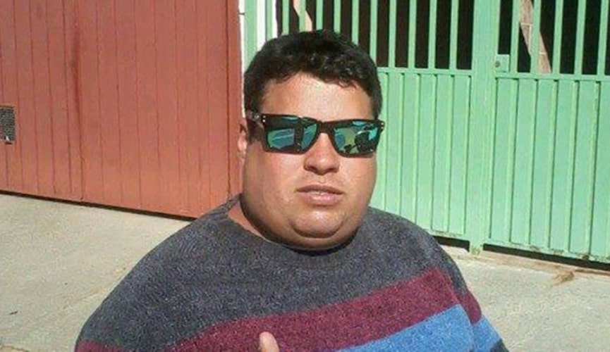 Homem com óculos de sol e camiseta cinza, vermelha e azul na frente de portão verde