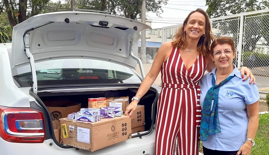 Carro com o porta-malas aberto, aparentemente, várias caixas de produtos de higiene; ao lado direito, uma senhora de cabelo curto com uniforme do hospital são vicente abraçada com uma mulher alta de macacão branco e vermelho listrado