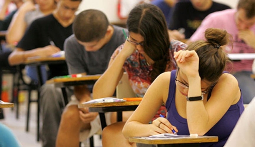 Mulher jovem loira e de óculos realizando prova na primeira cadeira, na segunda, outra mulher jovem com cabelos negros longos, na sequência rapaz pardo careca.