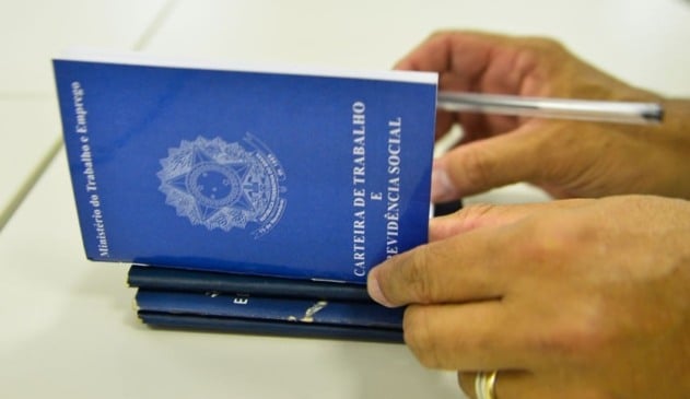 Carteira de trabalho sendo preenchida por mão de pessoa parda com aliança de casamento.