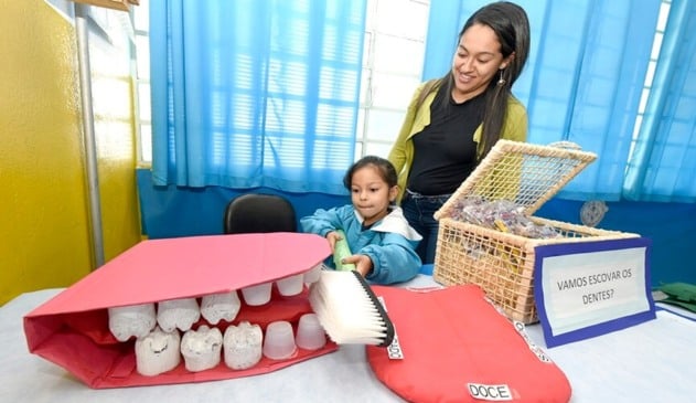 Menina escovando dentes de uma boca gigante de gesso, ao lado de uma mulher de cabelos escuros e com jaqueta verde