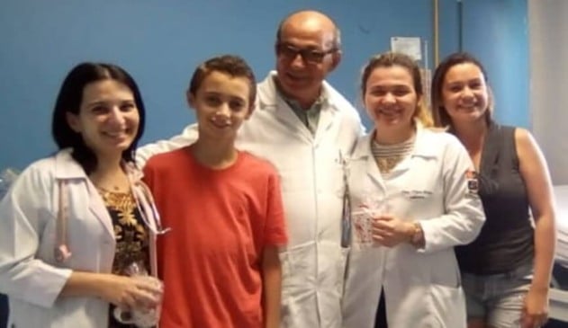 Menino de blusa vermelha ao lado de médicos e de mulher de blusa cinza