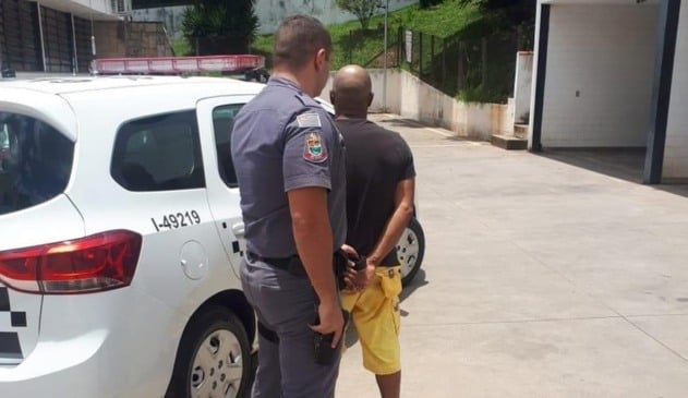 Homem sendo enquadrado por policial militar na frente de viatura branca da corporação