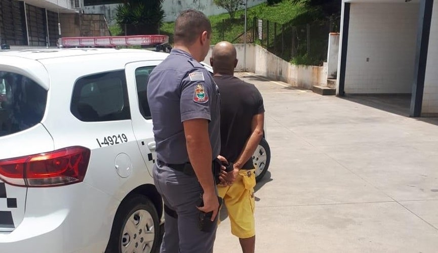 Homem sendo enquadrado por policial militar na frente de viatura branca da corporação