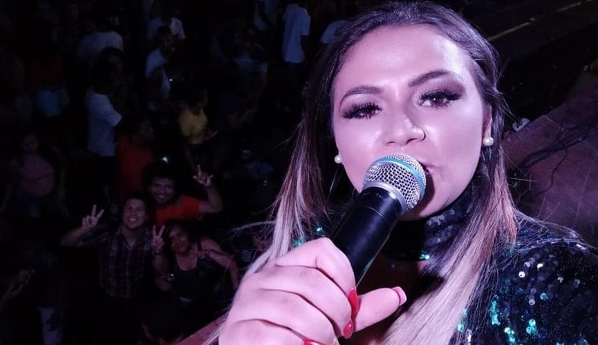 Selfie de mulher loira com microfone na boca, ao fundo pessoas