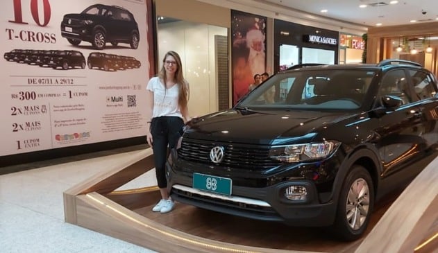 Mulher loira, com camiseta branca, calça preta e tênis branco, ao lado de um carro zero preto.