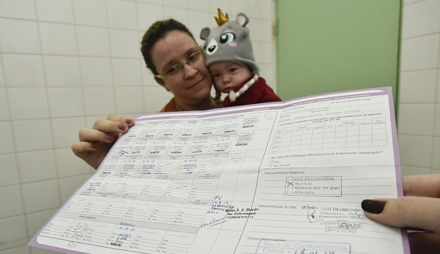 Mulher segurando criança com uma mão e com a outra a carteira de vacinação