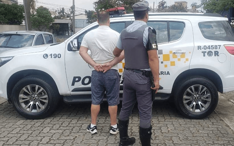 Imagem de policial segurando homem de costas, eles estão de frente para uma viatura da PR Imagem de policial segurando homem de costas, eles estão de frente para uma viatura da PR