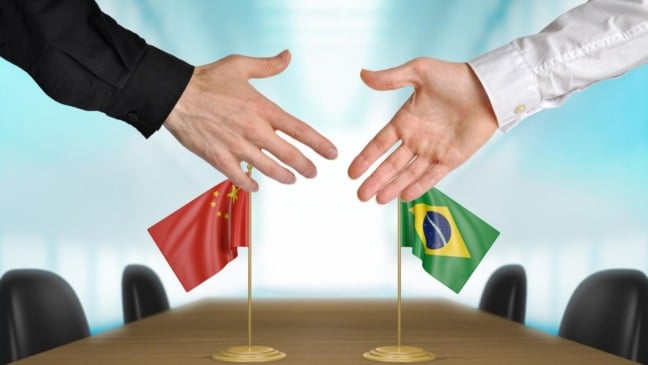 Dois homens iniciando um aperto de mãos com uma bandeira da China e uma bandeira do Brasil em cima de uma mesa