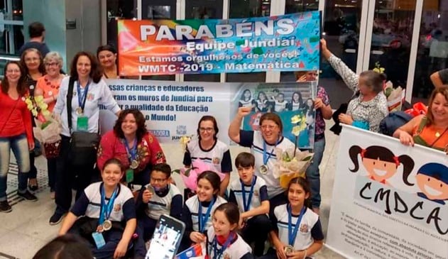 Alunos de jundiaí que participaram de olimpiada de matemática na china sendo repecionados ao chegar no brasil por familiares e professores. familiares seguram faixas parabenizando os alunos.