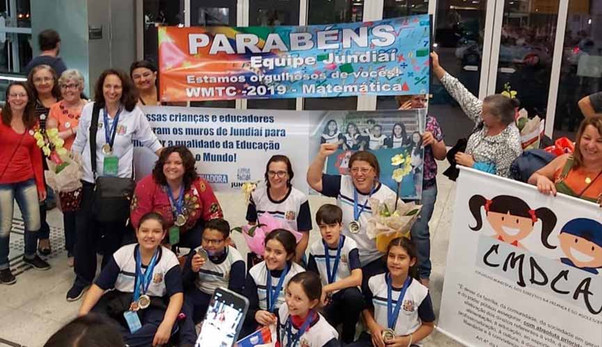 Alunos de jundiaí que participaram de olimpiada de matemática na china sendo repecionados ao chegar no brasil por familiares e professores. familiares seguram faixas parabenizando os alunos.