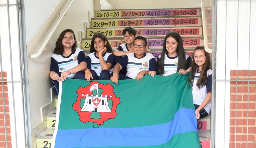 Seis crianças, sendo quatro meninas e dois meninos, sentados em escada com números da tabuada escritos e segurando a bandeira de Jundiaí