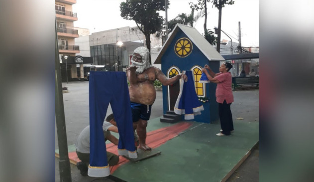 decoração de natal mostra papai noel negro e com samba canção pendurando vestes azuis em varal.. atrás, uma pequena casa azul de decoração e duas pessoas mexendo em detalhes da obra