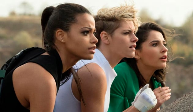 Reprodução de cena do filme As Panteras, em que as três personagens principais aparecem lado a lado e de perfil, em uma filmagem lateral, olhando para a frente atrás de um barranco