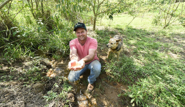 produtor rural mostra água limpa de nascente após trabalho de preservação. ele está em meio a mata, segurando um pouco da água. há um cachorro atrás dele