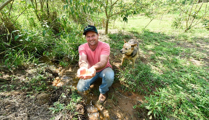 produtor rural mostra água limpa de nascente após trabalho de preservação. ele está em meio a mata, segurando um pouco da água. há um cachorro atrás dele