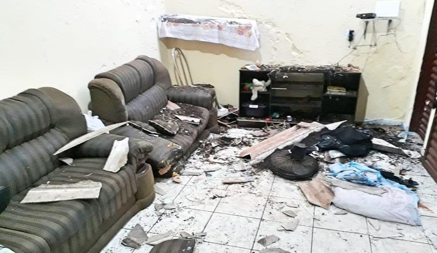 Televisão, ventilador, sofá e outros eletrodoméstico quebrados, junto aos estilhaços de telha do teto de uma sala