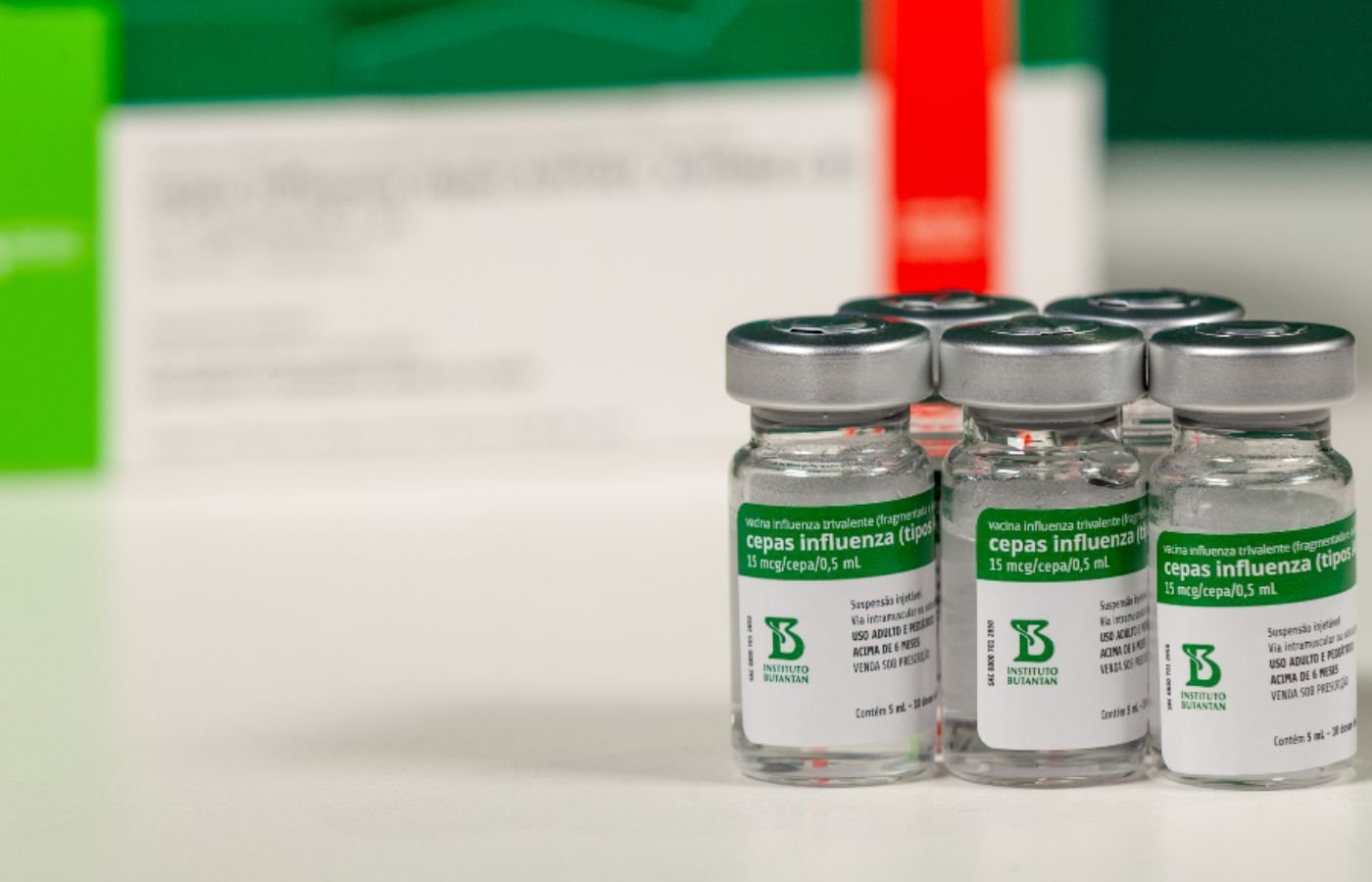 Frascos da vacina da gripe trivalente do Instituto Butantan organizados sobre mesa, com rótulo visível destacando cepas influenza.