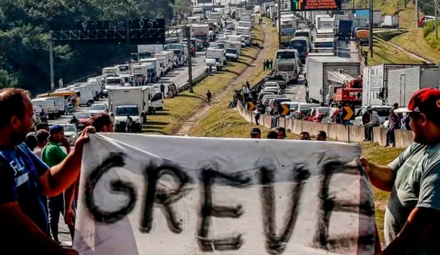 dois caminhoneiros seguram, cada um em uma ponta, uma faixa escrito "GREVE". Ao fundo, duas mãos de rodovia estão completamente tomadas por caminhões estacionados, durante greve de 2018