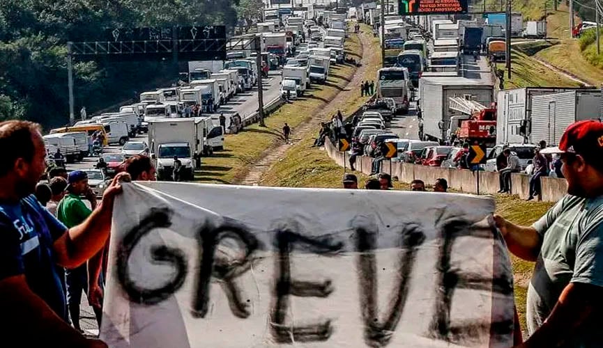 dois caminhoneiros seguram, cada um em uma ponta, uma faixa escrito "GREVE". Ao fundo, duas mãos de rodovia estão completamente tomadas por caminhões estacionados, durante greve de 2018