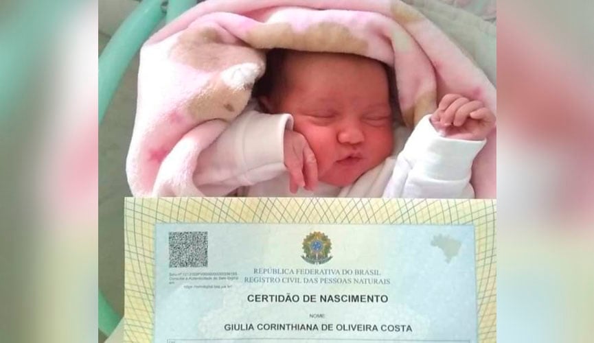 bebê deitada em cama dormindo com certidão de nascimento a sua frente, com o nome "giulia corinthiana de oliveira costa