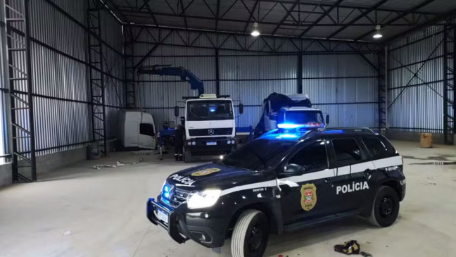 Galpão com caminhões desmontados e viatura da Polícia Civil com luzes ligadas em primeiro plano
