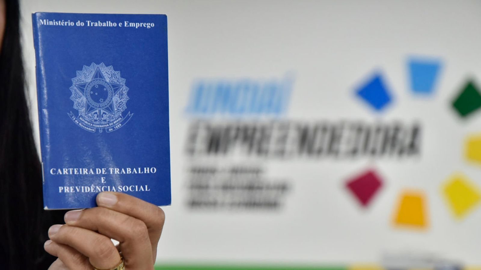 Carteira de trabalho sendo segurada à frente de parede da sede do Programa Jundiaí Empreendedora.