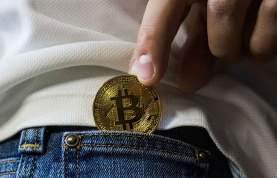 Pessoa colocando uma moeda física de Bitcoin no bolso da calça jeans.