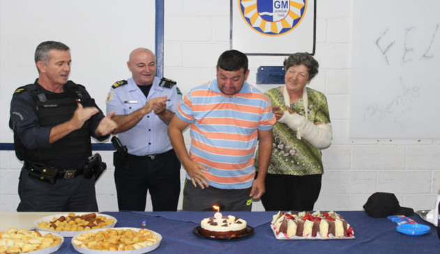 guardas municipais ao lado de bento e de sua mãe, com a mesa cheia de salgadinhos e com dois bolos, enquanto a vela em um bolo redondo e menor está acesa. os guardas batem palmas