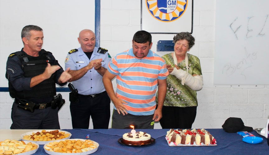 guardas municipais ao lado de bento e de sua mãe, com a mesa cheia de salgadinhos e com dois bolos, enquanto a vela em um bolo redondo e menor está acesa. os guardas batem palmas