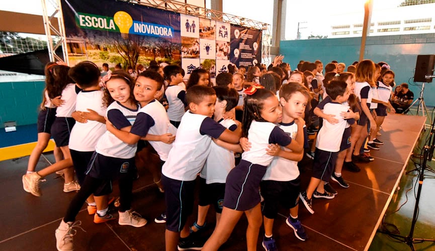 crianças fazem apresentação musical durante a entrega do espaço como Escola Inovadora. Eles estão de uniforme escola, em palco montado na quadra