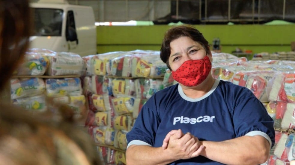 Mulher com uniforme da Plascar, em frente à doações de cestas básicas