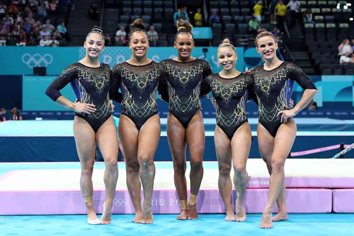 Equipe brasileira de ginástica artística posando na arena dos Jogos Olímpicos de Paris 2024, vestindo uniformes pretos com detalhes dourados.