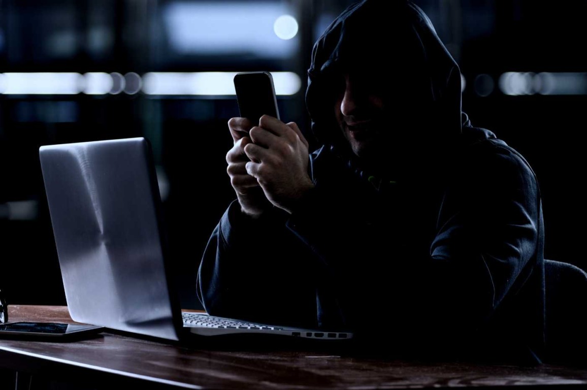 homem encapuzado em penumbra acessando o celular e notebook em atitude suspeita