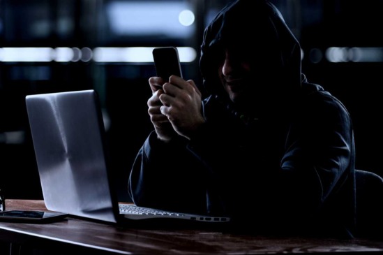 homem encapuzado em penumbra acessando o celular e notebook em atitude suspeita