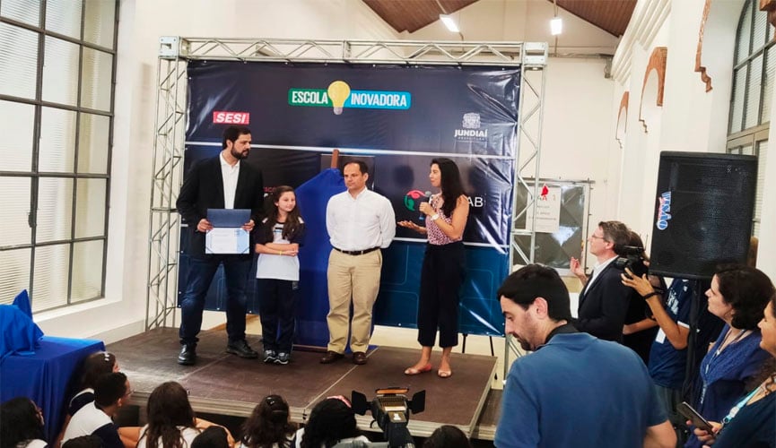 prefeito de jundiaí e 1º Diretor Secretário, Vandermir Francesconi Júnior das Federações das Indústrias de São Paulo na frente de painel da escola inovadora, segurando certificado e ao lado de duas mulheres. à frente, crianças e outras pessoas escutam sobre o anuncio do novo certificado do fablab