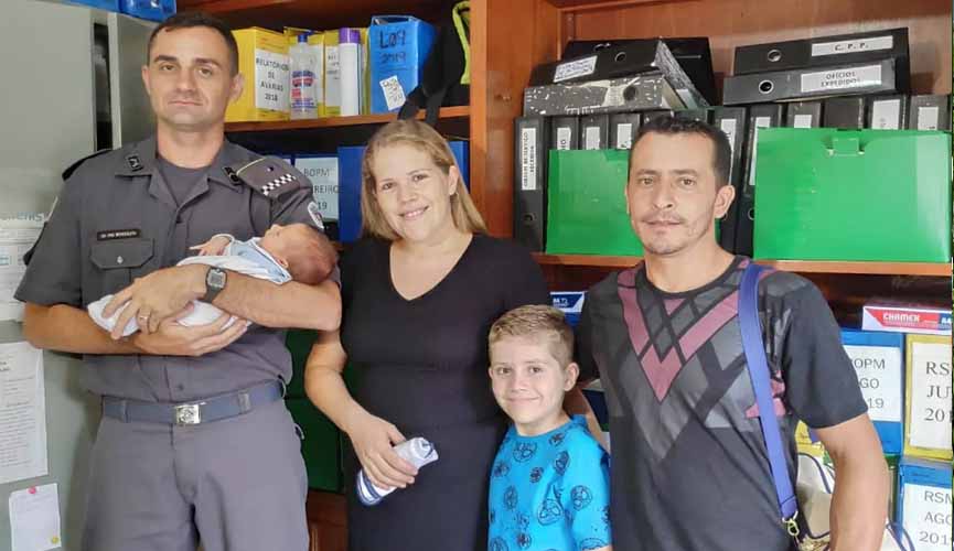 policial segura bebê no colo; ao lado está a mãe do bebê, com o outro filho menor ao lado e, depois, o pai