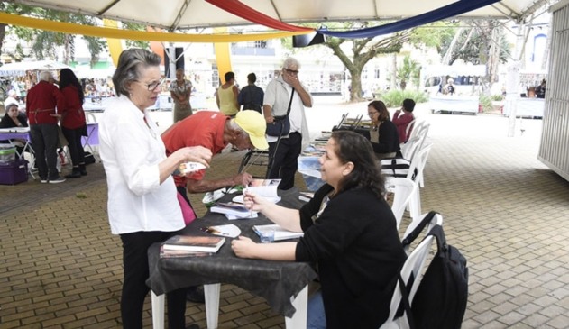 Mulher idosa com livro na mão com blusa branca, calça preta e óculos (à esquerda), de frente para mulher de meia idade com camisa preta e calça jeans sentada autografando livro na praça da Matriz de Jundiaí
