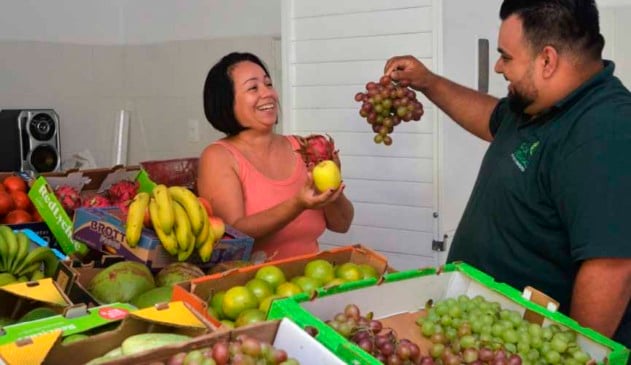 homem e mulher seguram frutas atrás de mesa cheia de caixotes com frutas e legumes