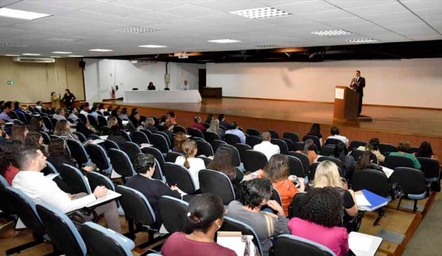 Pessoas em auditório sentadas na cadeira. No palco, à frente, homem fala