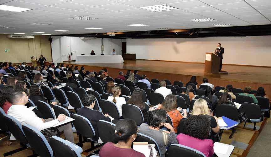 Pessoas em auditório sentadas na cadeira. No palco, à frente, homem fala